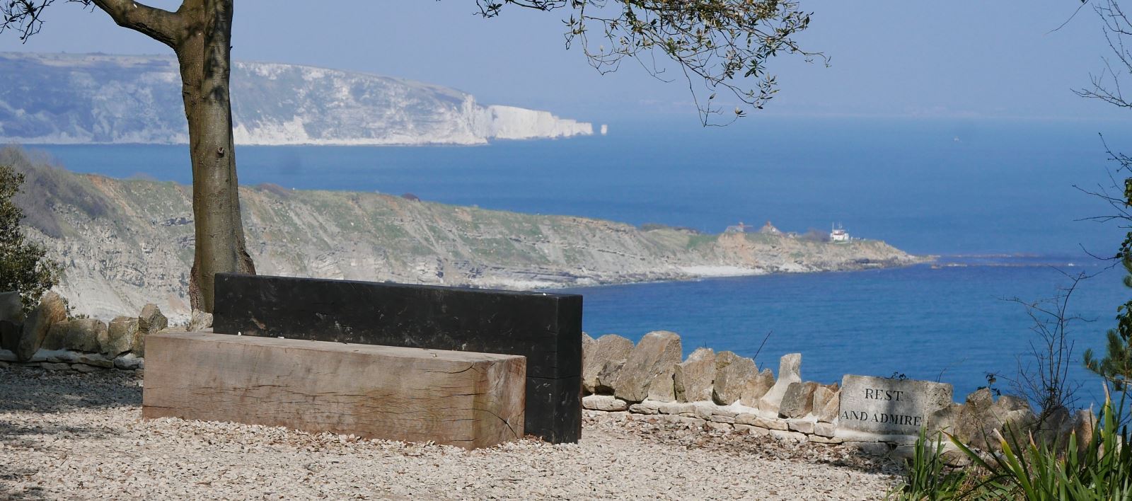A coastal view of two headlands Peveril Point and Ballard Down. Framed in the foreground by a tree wooden seat and a dry stone wall.